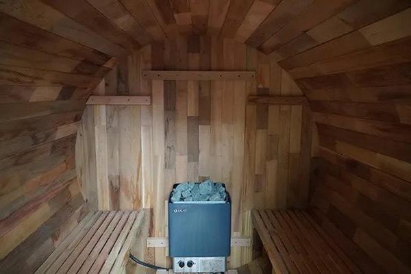 An indoor sauna made of wooden barrels with a rustic design. The sauna is filled with steam, and a person can be seen sitting inside, enjoying the warmth and relaxation. Wooden benches line the walls, and the lighting is soft and warm