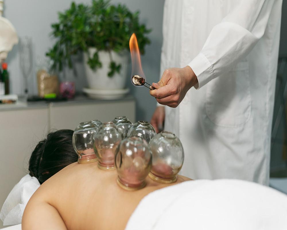 A close-up photograph of a person's hand holding a suction cup, while performing a massage technique on a client's back. The client is lying face down on a massage table, and the therapist's hand is visible in the foreground holding the suction cup against the client's skin