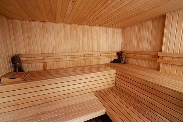 An interior photograph of a sauna room with wooden walls, ceiling, and benches. A stove is visible in the corner of the room with rocks on top of it. The lighting is dim and warm, creating a relaxing atmosphere