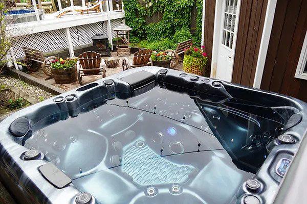 An outdoor hot tub surrounded by wooden decking with a wooden chair on one side and a lush green tree in the background